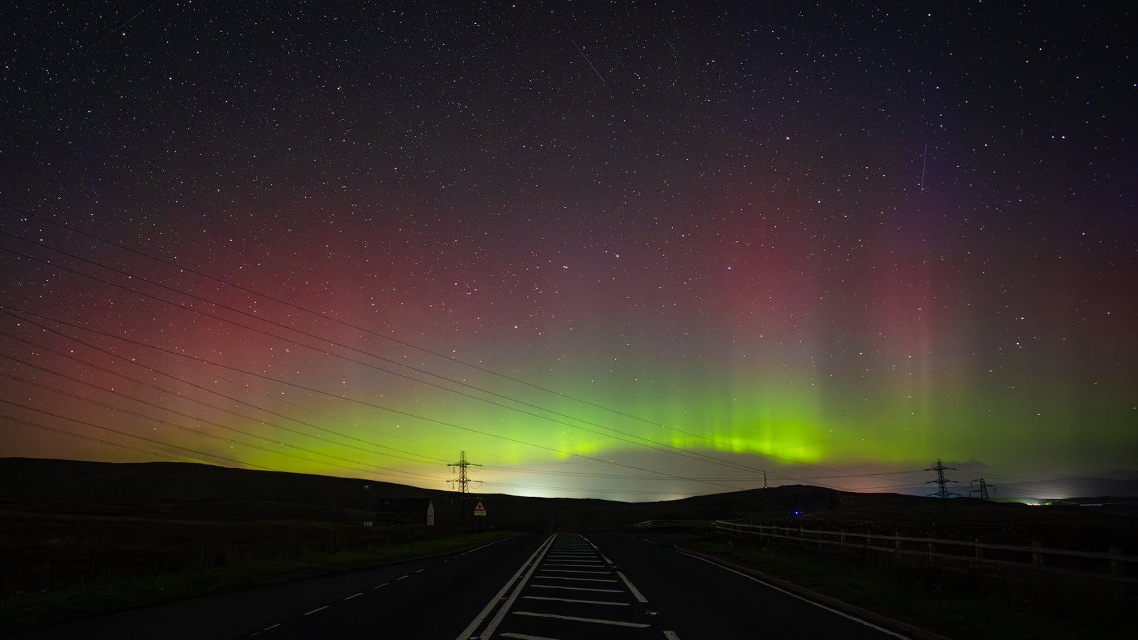 Green aurora rays with a red band above them, over a moorland road with pylons along the horizon.