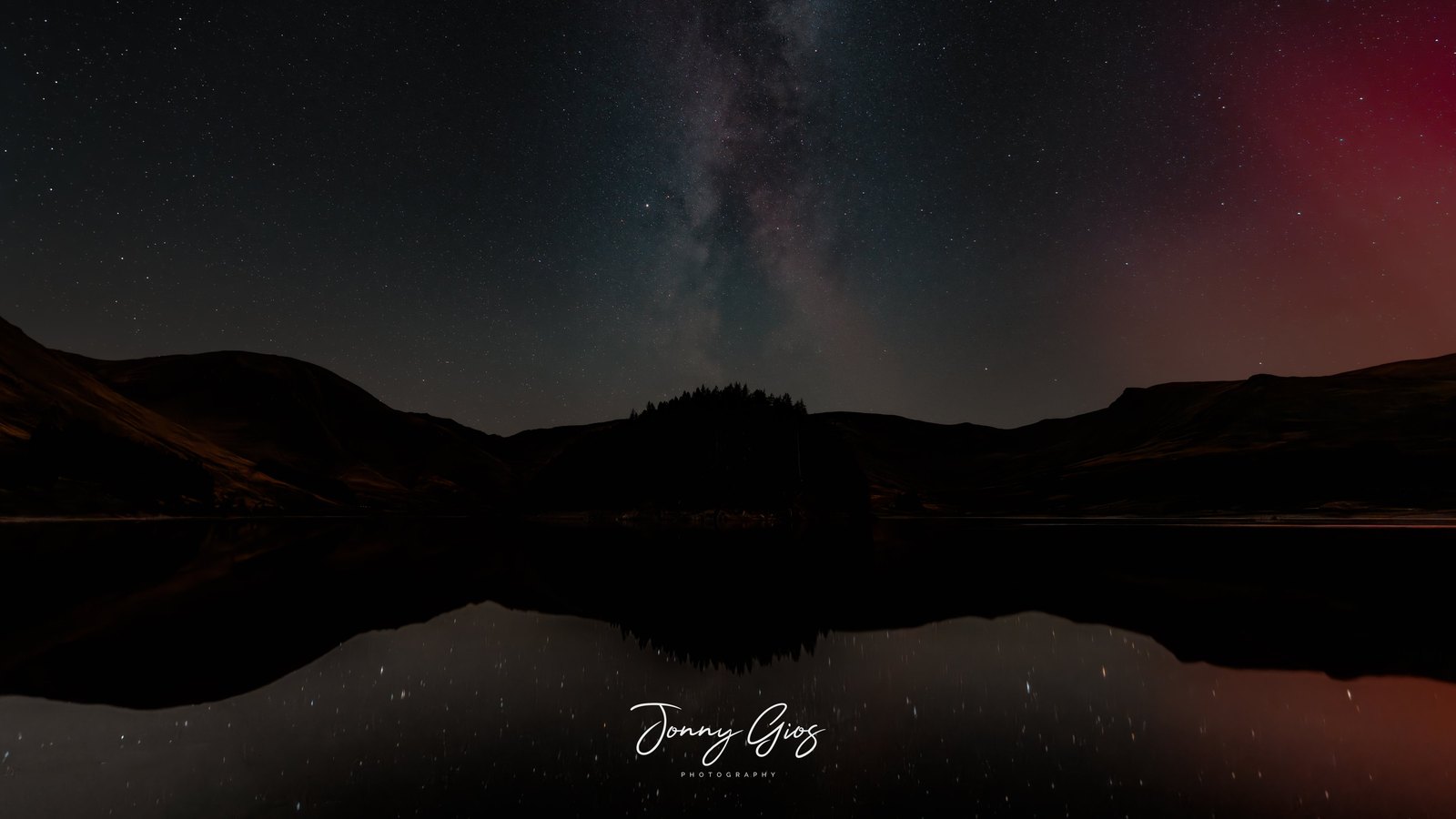 The Milky Way standing over a still lake, with a wooded island reflected in the water and a red aurora glow along the right of the sky.