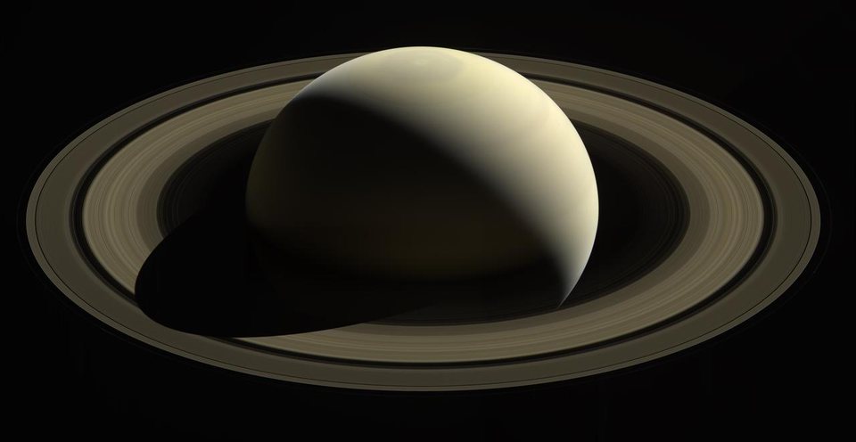 Saturn photographed by the Cassini spacecraft, the pale gold globe lit from one side and casting a shadow across its rings.